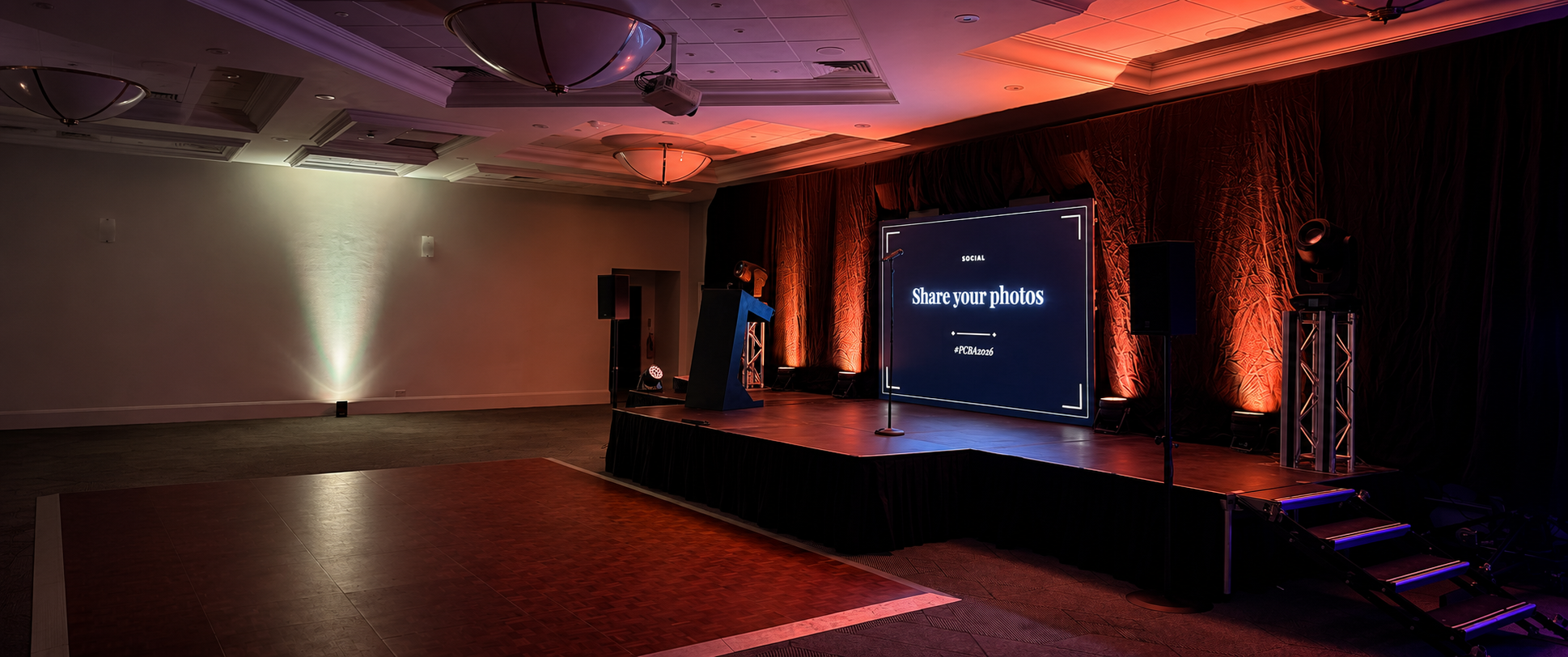 A large event hall with a raised stage, dark curtains, a central projection screen displaying 'Share your photos', a podium, speakers, and a dance floor under warm and cool stage lighting.